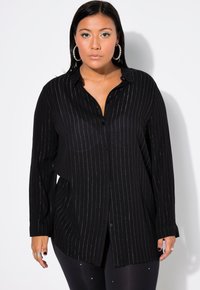 Schwarze Hemdbluse mit vertikalen silbernen Streifen; lange Ärmel, Kragen und lockere Passform. Kombiniert mit schwarzen Leggings mit kleinen Akzenten.