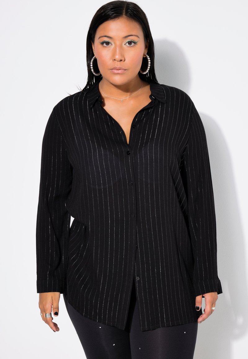 Schwarze Hemdbluse mit vertikalen silbernen Streifen; lange Ärmel, Kragen und lockere Passform. Kombiniert mit schwarzen Leggings mit kleinen Akzenten.