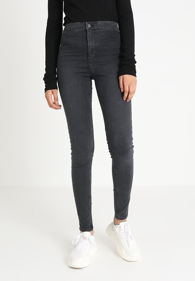 Žena oblečená v tmavě šedých skinny jeans s vysokým pasem, černém dlouhém tričku s dlouhým rukávem a bílých robustních teniskách, stojící před jednoduchým bílým pozadím.