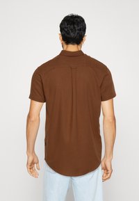 Camicia marrone a maniche corte in tessuto testurizzato, con piega posteriore, colletto e orlo arrotondato. Nessun logo o decorazione visibile.