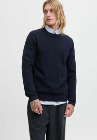 Jersey de cuello redondo en color navy, confeccionado en un tejido suave, con puños y dobladillo de canalé. Llevado sobre una camisa de rayas azul claro, complementado con unos pantalones grises.