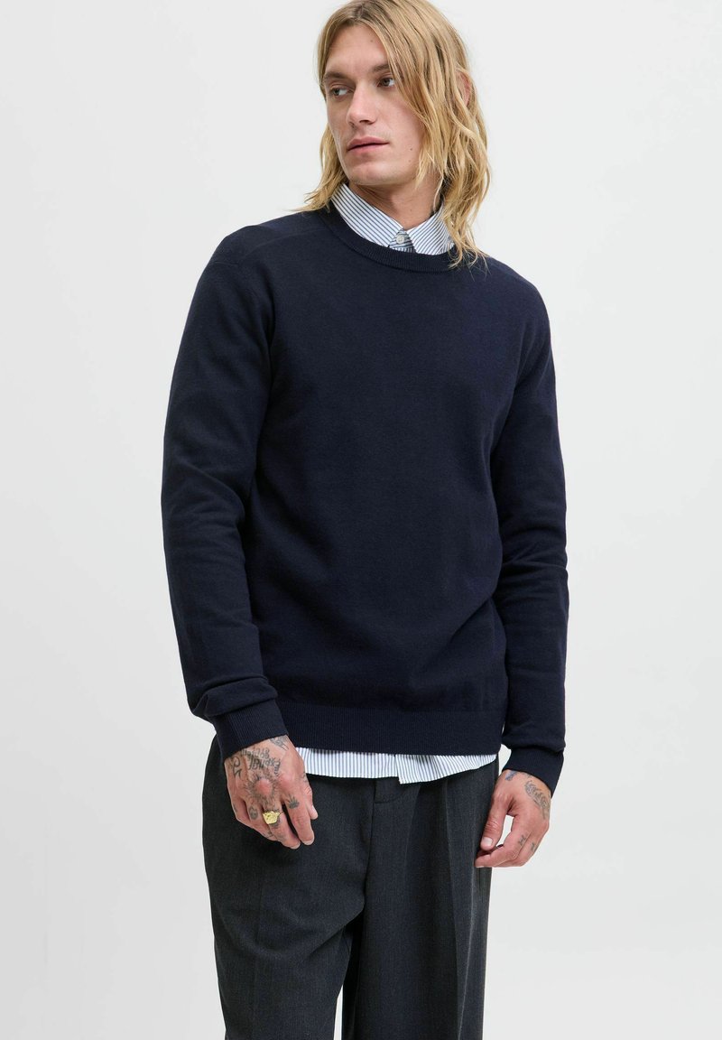 Jersey de cuello redondo en color navy, confeccionado en un tejido suave, con puños y dobladillo de canalé. Llevado sobre una camisa de rayas azul claro, complementado con unos pantalones grises.