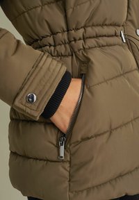Olive green puffer jacket with a zippered pocket, elastic ribbed cuffs, and horizontal stitching; features a metallic snap button detail.