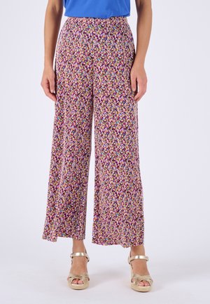 Mujer con pantalones de pierna ancha con un estampado floral colorido y sandalias metálicas doradas con tiras en el tobillo, de pie contra un fondo liso.