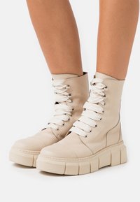Botas altas bege com sola de borracha grossa, apresentando atacadores brancos e ilhós prateados. Superfície de couro liso com acabamento cosido.