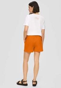 Witte T-shirt met oranje detail op de rug, gecombineerd met oranje shorts. De outfit heeft een elastische tailleband en een casual pasvorm. Zwarte sandalen maken de look compleet.