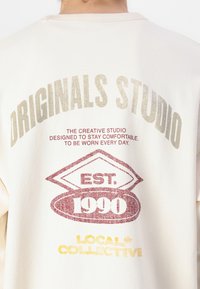 Sudadera color crema con texto gráfico en negrita en dorado, burdeos y amarillo. El texto incluye "Originals Studio" y "EST. 1990."