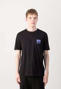 T-shirt en coton noir avec un patch rectangulaire violet sur la poitrine gauche portant le texte "HUGO". Manches courtes, col rond, tissu uni.
