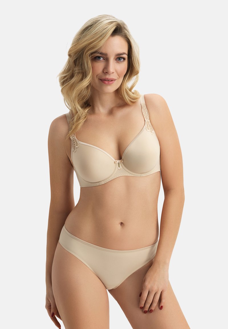 Mujer con cabello rubio ondulado, vistiendo un conjunto de sujetador y bragas beige a juego, de pie frente a un fondo claro y liso.