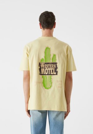 Gul bomull t-skjorte med grønn kaktusgrafikk og "WESTERN MOTEL" tekst på baksiden. Løs passform, korte ermer, uformelt design.
