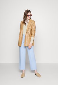 Brun läder dubbelknäppt blazer, ljusgrön topp, ljusblå vida byxor och beige flätade slipskor; klara rektangulära solglasögon.