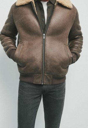 Mężczyzna w brązowej skórzanej kurtce bomber z kołnierzem z owczej wełny, ręce w kieszeniach, w ciemnoszarych dżinsach i zielonej koszuli pod spodem.