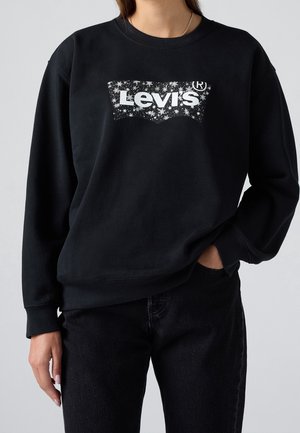 Personne portant un sweat-shirt Levi's noir avec logo étoile blanche, associé à un jean noir, une main dans la poche sur fond uni.