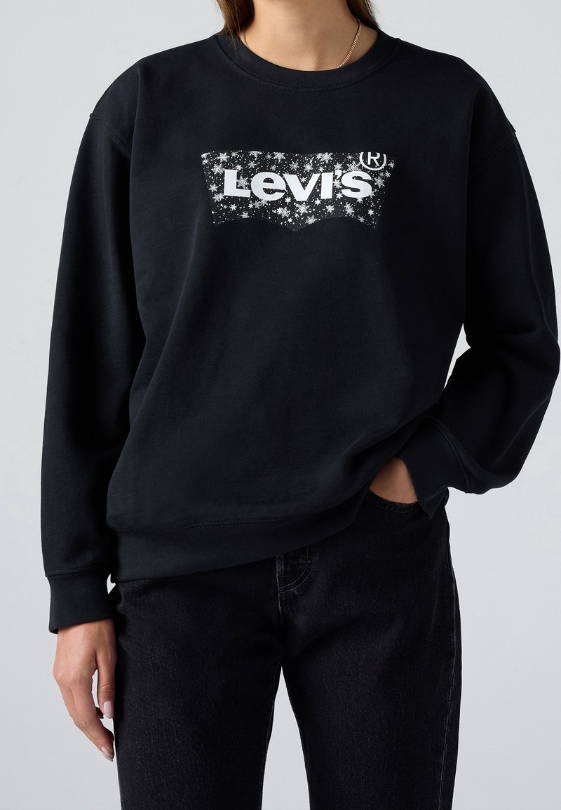 Personne portant un sweat-shirt Levi's noir avec logo étoile blanche, associé à un jean noir, une main dans la poche sur fond uni.