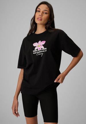 Kobieta stoi z ręką na biodrze, ubrana w czarny T-shirt z różowym kwiatem i napisem oraz czarne prążkowane szorty na jednolitym tle.
