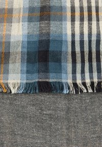 Tissu à carreaux dans des tons de bleu et de blanc avec des accents noirs, présentant une bordure à franges. En dessous, un tissu texturé gris.