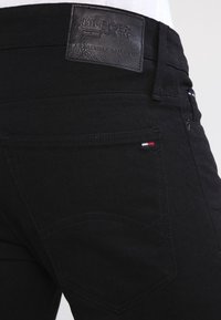 Black denim jeans feature a leather label at the waistband with embossed text, subtle stitching, and a small flag color accent on the back pocket.