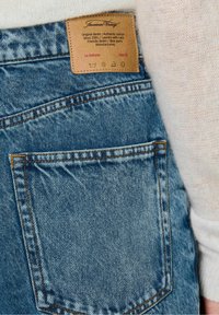 Blauwe denim jeans met een lichte wassing, voorzien van een rechthoekige achterzak en een gemerkte tan leren patch erboven. Contraststiksels zijn zichtbaar.