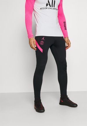 Persoon draagt zwarte en roze trainingsbroek van Paris Saint-Germain, wit-roze longsleeve en zwarte sportschoenen met rode accenten.