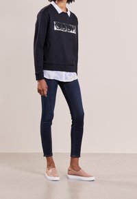 Osoba ubrana w czarną bluzę z błyszczącym logo, białą koszulę z kołnierzykiem, ciemne obcisłe dżinsy oraz beżowe trampki na slip-on, stoi na neutralnej podłodze.