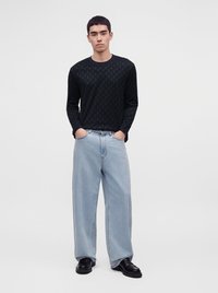 Giovane uomo in piedi con le mani nelle tasche, indossa una camicia nera a maniche lunghe con motivo, jeans azzurri a gamba larga e scarpe nere su sfondo bianco.