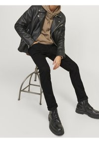 Persoon gekleed in een zwarte leren jas, beige hoodie, zwarte broek en zwarte laarzen, zittend op een metalen kruk tegen een effen achtergrond.