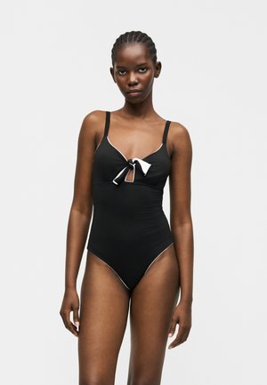 Kobieta w czarnym jednoczęściowym kostiumie kąpielowym z białą lamówką i kokardą z przodu, stojąca na jasnym jednolitym tle.