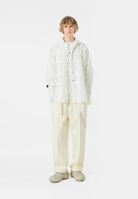 Camisa blanca de gran tamaño con detalles de parches rectangulares, combinada con pantalones amarillos claros y calzado gris texturizado. Diseño simple y colores apagados.