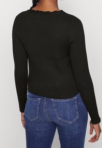 Top lungo a maniche nere a coste con scollatura a ondas, abbinato a jeans di denim blu con dettagli sulle tasche posteriori.