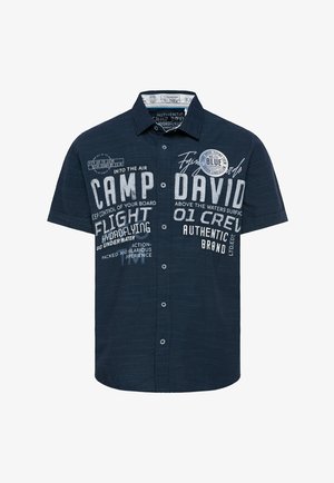 Rövid ujjú, gombos ing tengerészkék színben, texturált felülettel. Jellegzetes fehér szöveges grafikákkal és különféle nyomott díszítésekkel rendelkezik.