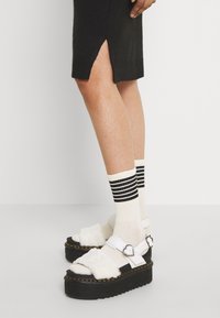 Bílé platformové sandály s umělou kožešinou a stříbrnou srdcovou sponou, doplněné krémovými ponožkami s černými vodorovnými pruhy a černými pletenými šaty.