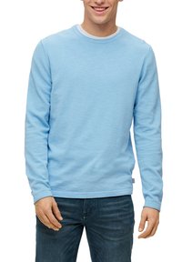 Young man smiling, wearing a light blue long-sleeve shirt and dark blue jeans, standing against a white background.