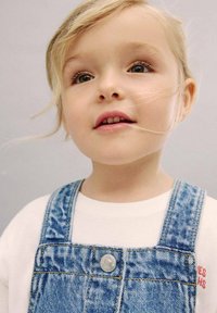 Džínové kousky s nastavovacími popruhy, stříbrným knoflíkem, v kombinaci s bílou košilí s červeným textem, na jednobarevném pozadí.