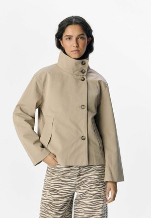 Frau trägt eine beige Jacke mit hohem Kragen und Knöpfen sowie Zebrahosen, steht mit einer Hand in der Tasche vor einem schlichten Hintergrund.