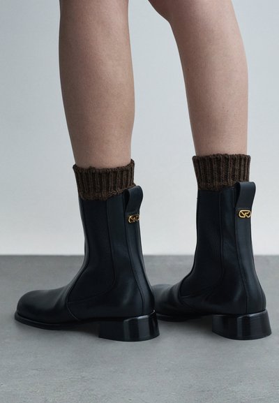Czarne skórzane botki za kostkę o strukturalnym wzorze, brązowe prążkowane mankiety z dzianiny i złote detale z tyłu. Niskie obcasy na klocku.