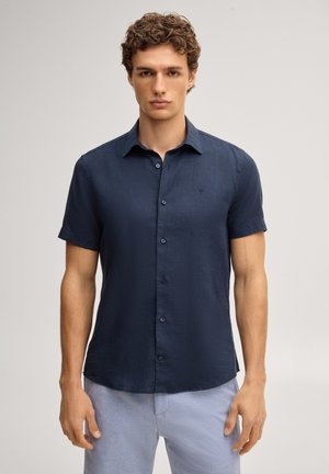 Mężczyzna ubrany w dopasowaną granatową, krótką koszulę na guziki i jasnoniebieskie spodnie z teksturą, stojący na jednolitym tle.