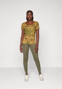 Olivgrön tie-dye t-shirt med kort ärm i kombination med olivgröna leggings. Modellen bär vita sportskor. Enkel, åtsittande design.