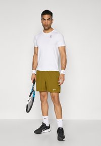 Mann in weißem Sportshirt, olivgrünen Shorts, weißen Handgelenkbanden und Socken, schwarzen Sneakers, der einen Tennisschläger hält.
