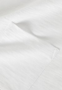 Tessuto bianco strutturato con cucitura in evidenza e dettagli di tasca. Il materiale appare morbido e leggero con sottili variazioni nella trama.