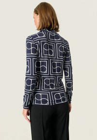 Marineblå mønstret langærmet top med geometriske hvide design. Glat tekstur med en tætsiddende pasform, båret med sorte bukser.