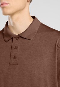 Polo shirt marron avec un col côtelé et une patte de boutonnage à trois boutons. Le tissu a une texture lisse avec un motif chiné subtil.