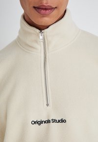 Cream-colored fleece quarter-zip top featuring a silver zipper and "Originals Studio" embroidered in black at the chest.
