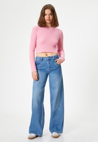 Top corto ribbed rosa con maniche lunghe, abbinato a jeans a gamba larga blu a vita alta. Calzature in bianco con accenti beige.