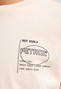 Kurzärmliges, hellbeiges T-Shirt aus Baumwolle. Verfügt über ein bedrucktes schwarzes Etikett mit "PETROL" und zusätzlichen Texten in verschiedenen Schriftarten.