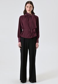 Blusa bordeaux con colletto a fiocco e scollo arricciato, abbinata a pantaloni neri a gamba larga. Tessuto liscio e maniche lunghe con polsini a bottone.