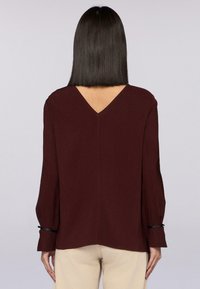 Donna con capelli neri e lisci che indossa un top a maniche lunghe bordeaux con scollo a V sulla schiena e pantaloni beige, in piedi con la schiena visibile.