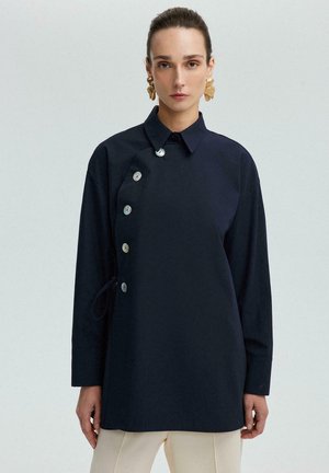 Camicia blu navy con colletto, maniche lunghe e chiusura a bottoni sul lato sinistro. Il tessuto è liscio con un design strutturato.