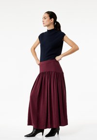 Top sin mangas azul marino, de cuello alto, combinado con una falda maxi de burdeos profundo, fruncida en la cintura. Botines negros de tacón alto completan el atuendo.