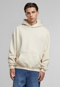 Felpa con cappuccio color crema, dotata di tasca frontale, polsini elastici e un logo discreto sul petto, abbinata a jeans blu.