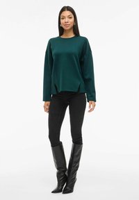 Sudadera oversized de color verde oscuro con mangas largas y aberturas laterales, combinada con unos jeans skinny negros y botas altas de cuero negras con punta. 